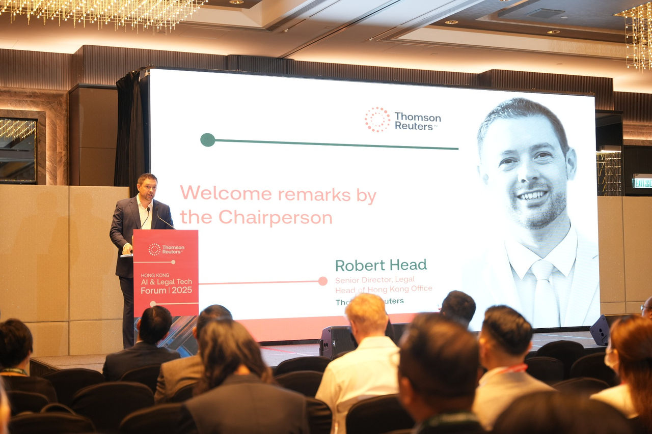 Chairperson delivering welcome remarks onstage with a large presentation screen behind, addressing an audience at the Hong Kong AI & Legal Tech Forum.